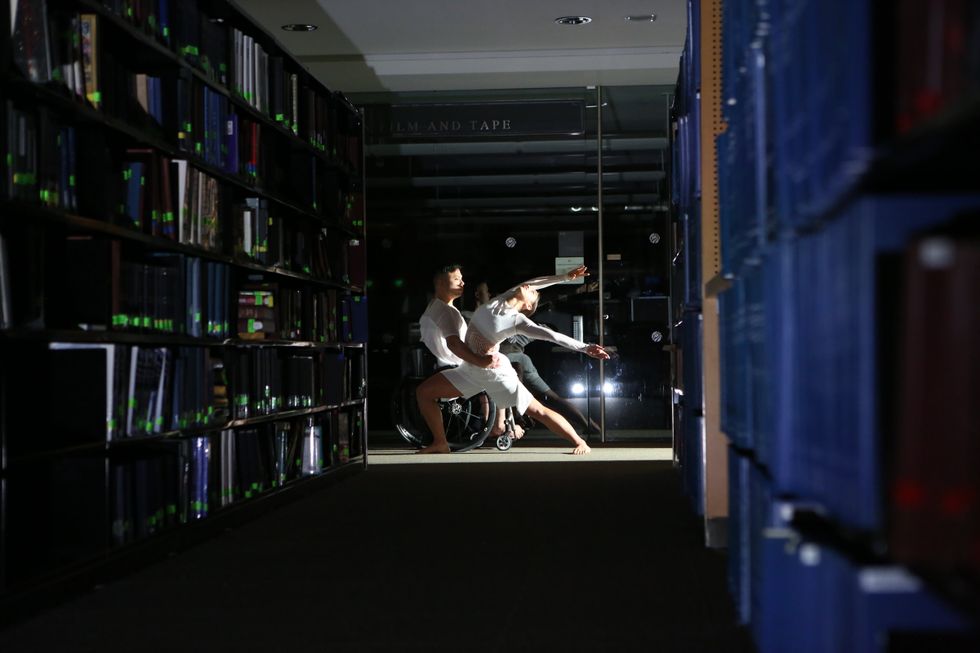 At the end of a darkened aisle of library books, a male dancer in a wheelchair curves an arm around the waist of a female dancer, who stands in a deep lunge as she arches backwards, arms rising around her head.