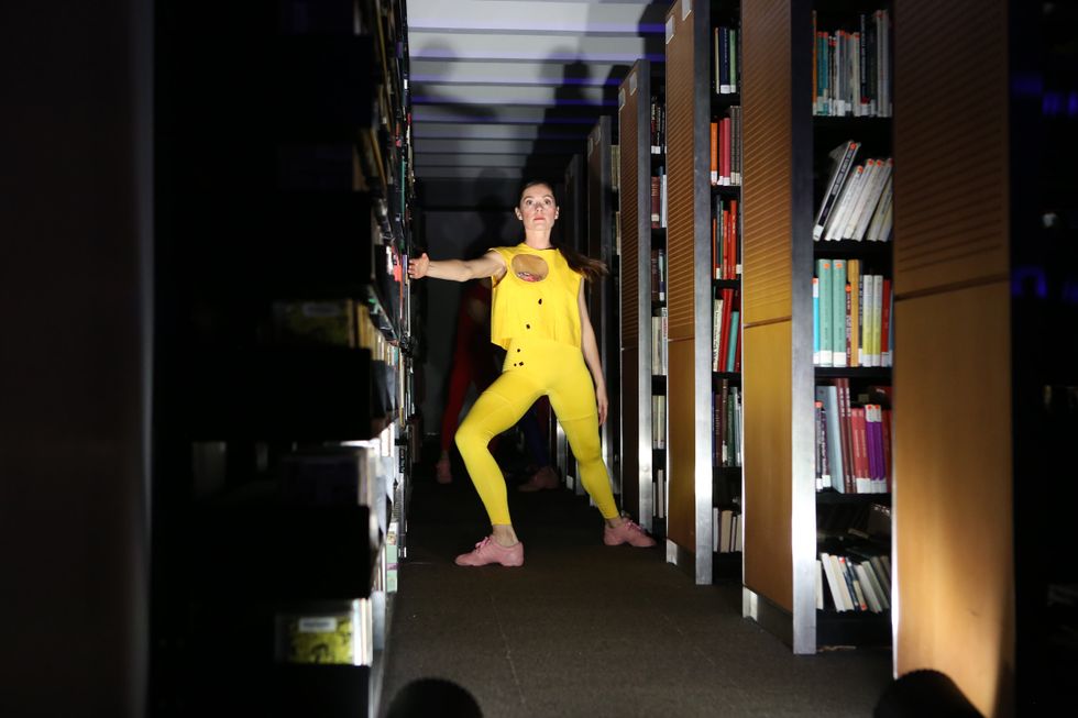 A woman in bright yellow tights and a matching, boxy shirt lunges, facing the camera. Her extended right arm touches the shelves to the left; her turned out back foot brushes the edge of the shelves to the right. The lighting is bright in the aisle in which she stands but shadowy elsewhere.