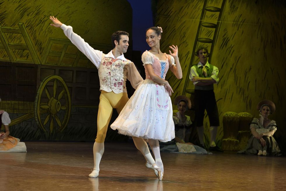 u200bLetizia Galloni, a light-skinned Black woman, balances in passu00e9 derriu00e8re on pointe while smiling at her partner, Mathias Heymann, a white male with dark hair. He stands behind her in tendu, his hand on her waist.