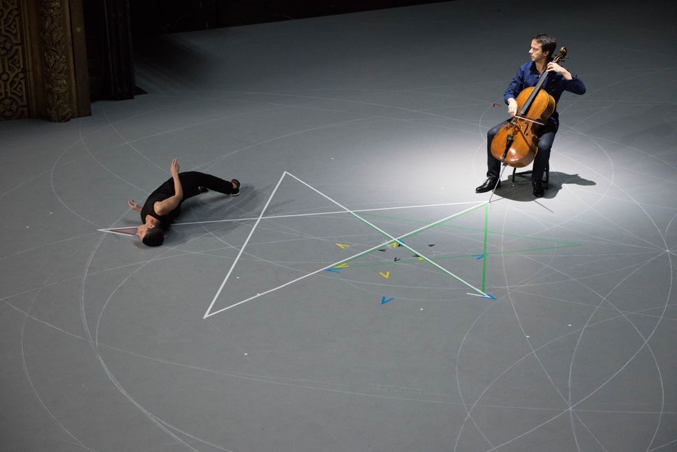 On a broad, grey stage, a cellist watches a woman in black arching on her side on the floor, partially covering a five-pointed star made of tape.