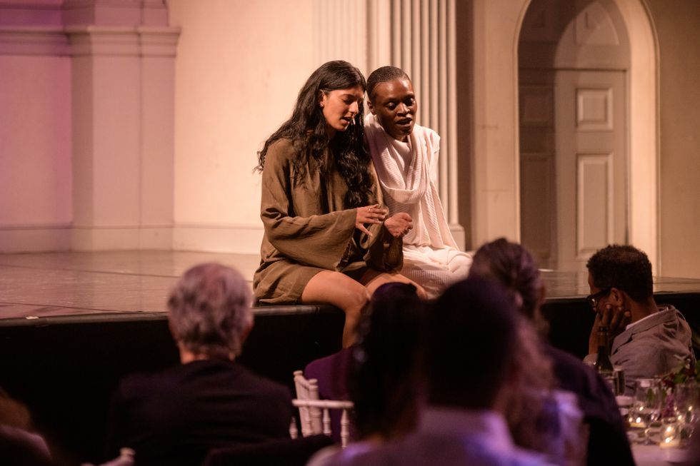Samita Sinha and Okwui Okpokwasili sit with their legs dangling off the edge of a stage in a well-lit room. They are shoulder to shoulder, eyes downcast as they open their mouths, as though singing.