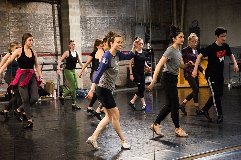 Michelle Dorrance smiles at the mirror, where a studio full of dancers is reflected as they imitate her movement.