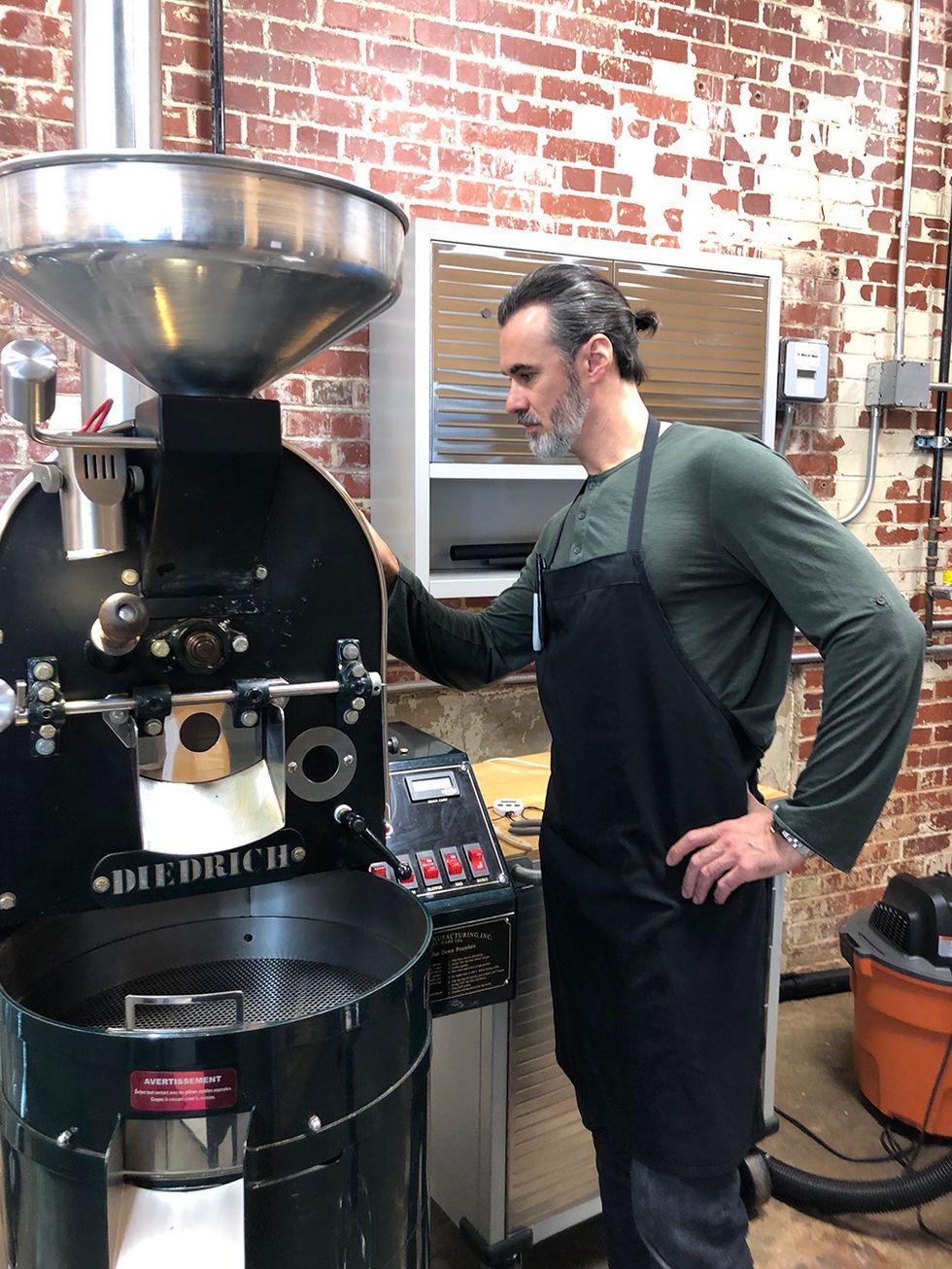 Paul, wearing a black apron, stands next to a large machine used to roast or blend the coffee.