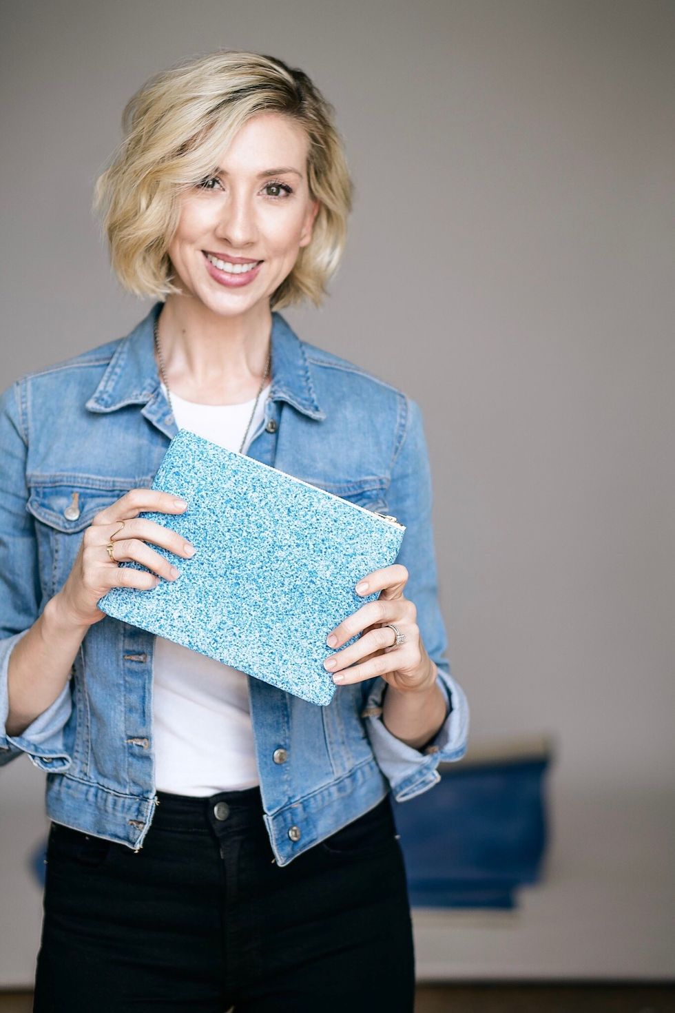 Founder Jennifer Kahn poses with a speckled blue clutch. She has wavy blonde hair and is wearing a denim jacket, white shirt and black pants.