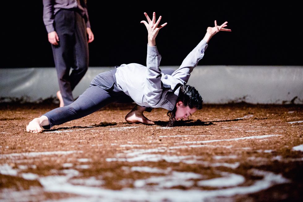 A dancer lunges down with her face almost next to a stage covered in dirt, her fingers splayed as her arms reach behind her