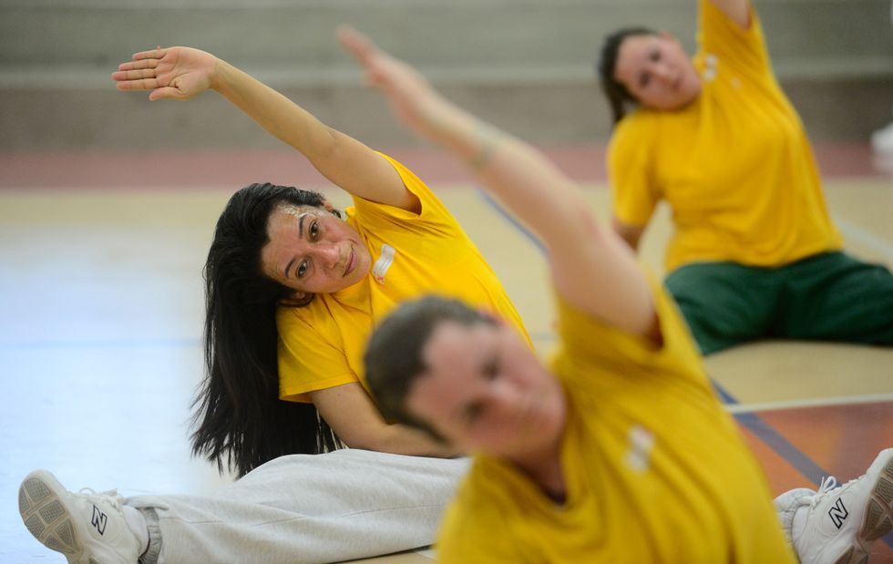 A group of women in prison, wearing yellow T-shirts and gray sweatpants, are stretching on a gym floor in a straddle split with one arm reaching to the side.
