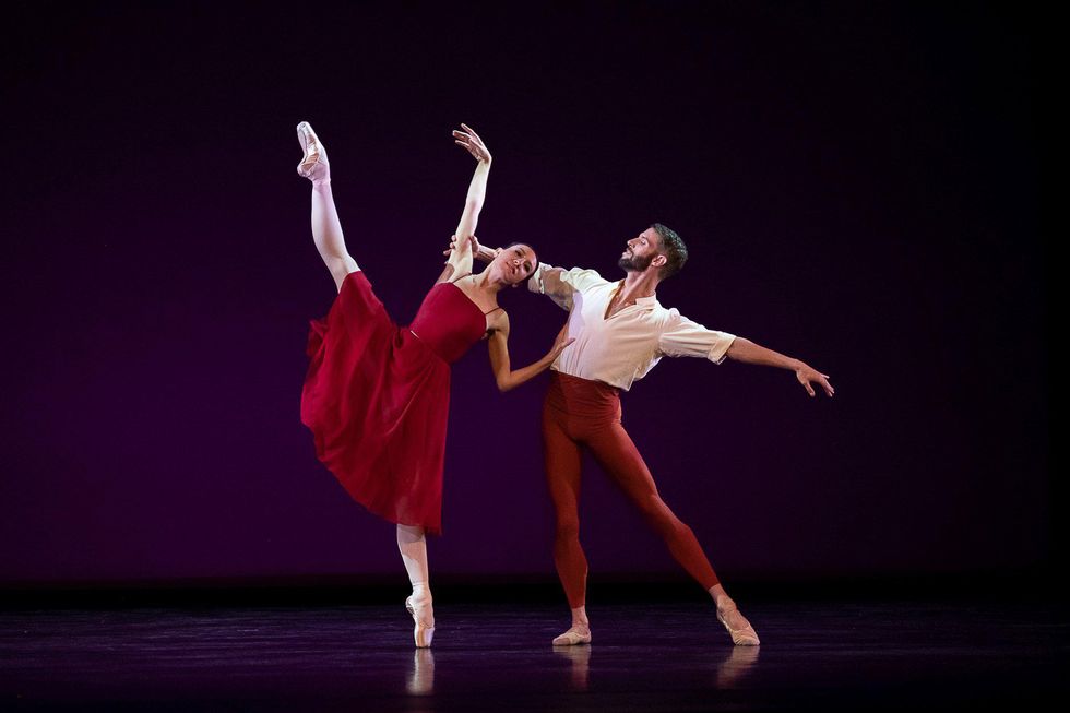Isabella Boylston, in a red dress, developpes to the side while leaning toward James Whiteside, who stands in tendu side.