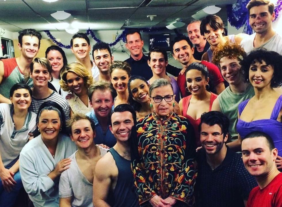 A solemn Ruth Bader Ginsburg, dressed in a colorful blouse and bright green earrings, stands surrounded by smiling young cast members, still in show makeup.