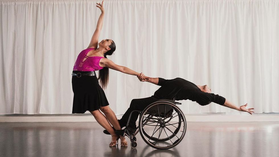 Marisa Hamamoto partners with Piotr Iwanicki, in a wheelchair. She's standing in heels, holding his hand and they both counterbalance each other and lean away