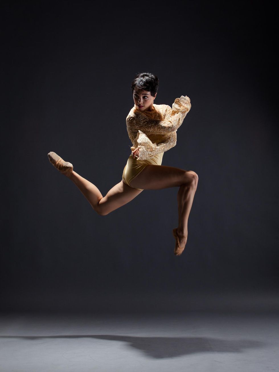 Allison Walsh jumps high on a grey background, legs split in either direction and bent at 90 degree angles