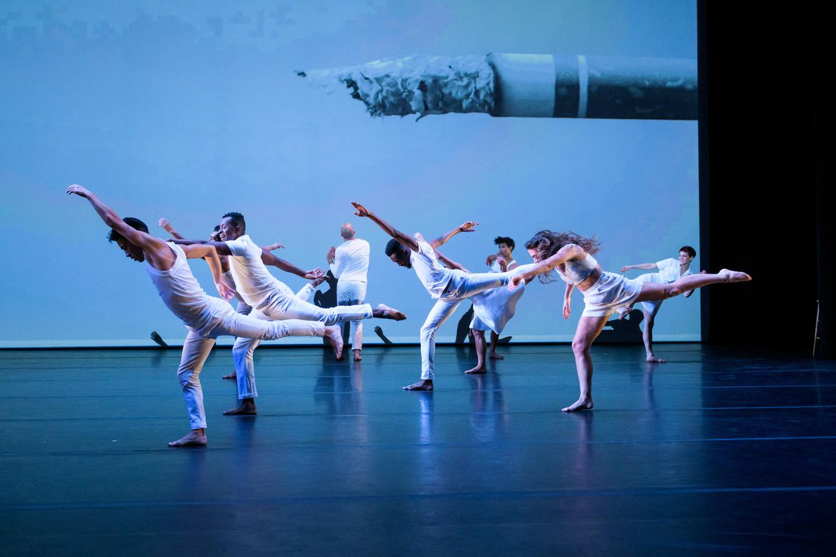 Several dancers are moving on stage, while dressed in all-white costumes. The five most downstage have their left arms and legs stretched in front and behind them. They are supported by their right leg which is slight bent. Projected behind them on the backdrop is a partially used cigarette.