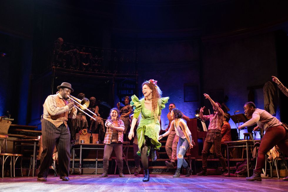 Amber Gray dances in a green dress, while a man plays trombone. The Workers Chorus dances behind her.