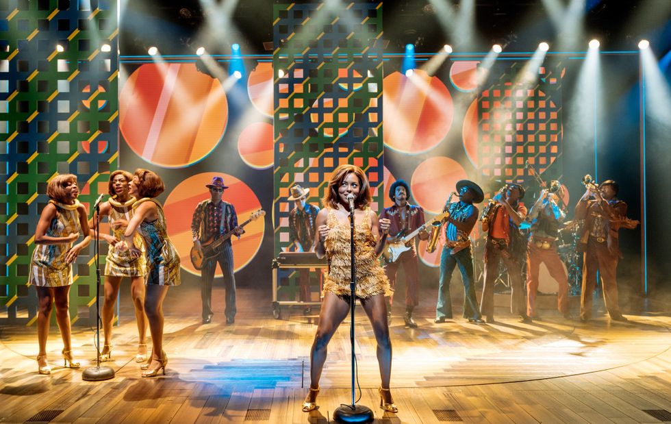 A production still from Tina. Tina, in a short gold dress, sings, flanked by a band to her right and backup singers to her left.
