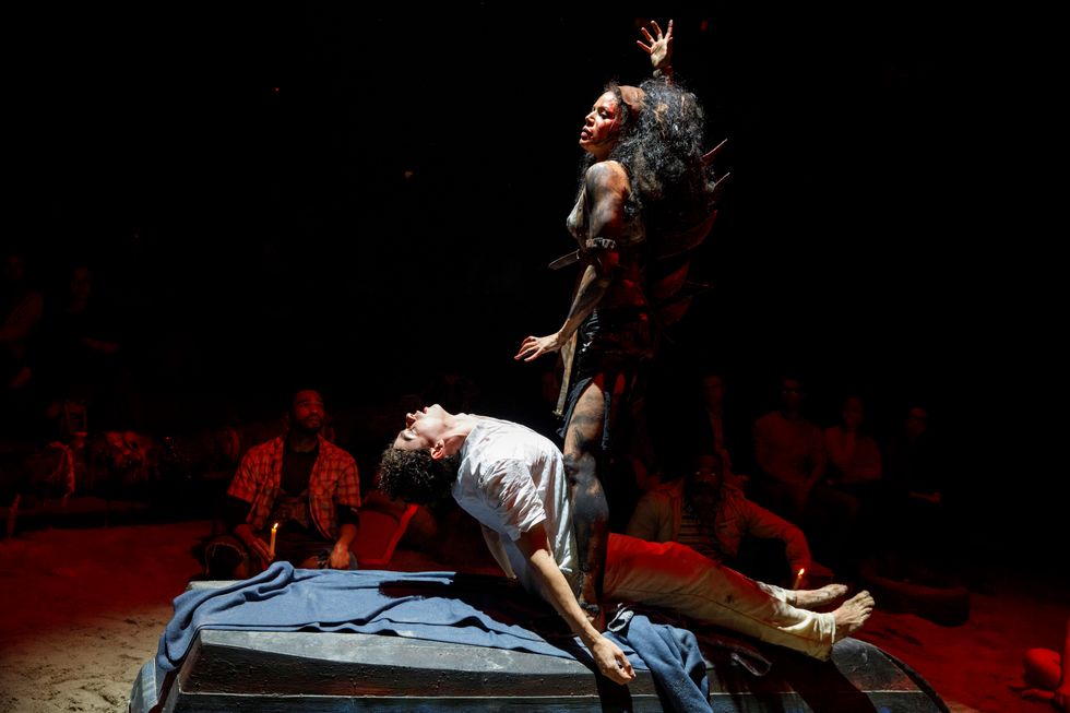A man lies on a bed, his chest arched up as if being summoned by the woman standing above him. She has paint on her body and disheveled hair and motions as if she is cursing him. The audience can be seen dimly in the background. 