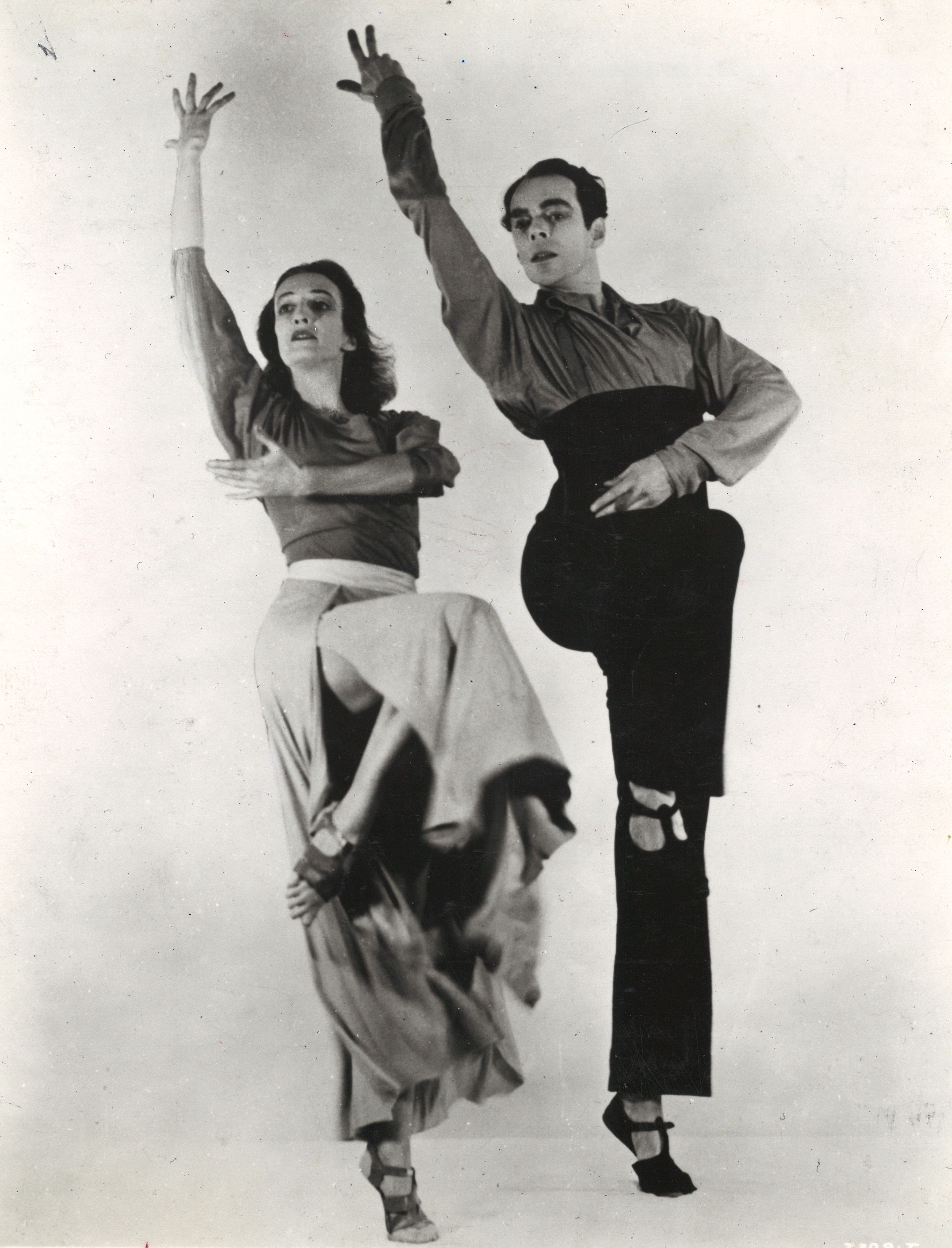 Humphrey and Weidman pose on relevu00e9, working legs raised to parallel retiru00e9, one arm raised overhead and slightly behind, opposite arms wrapping across their torsos