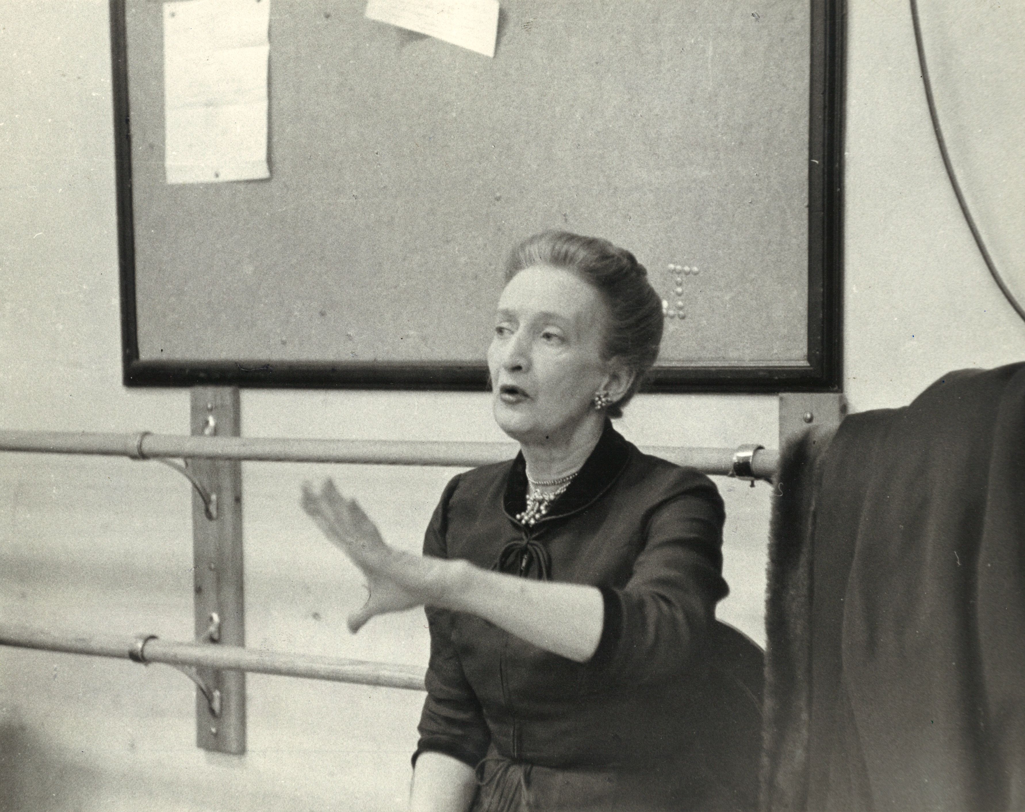 An older Doris Humphrey, wearing a high-necked blouse with pearls, hair elegantly pulled back, is caught mid-speech, gesturing with one hand as she sits in front of wall barres.