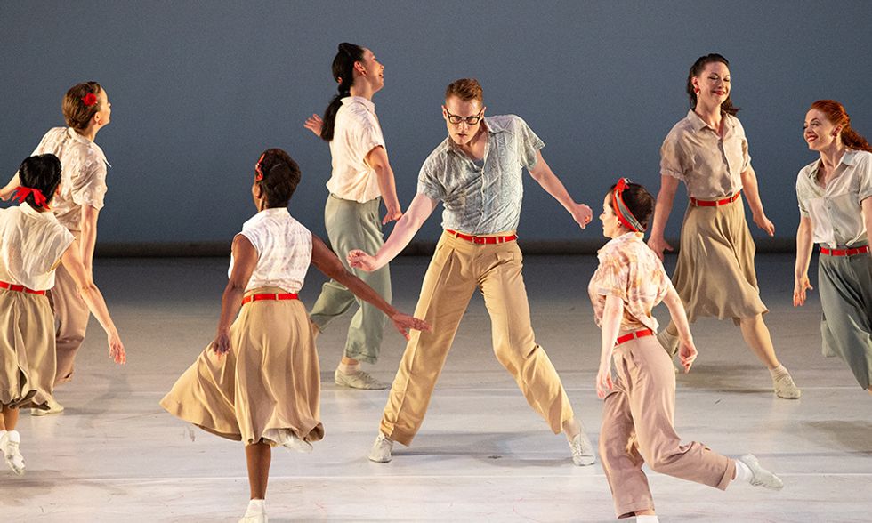 A male dancer in khakis, a red belt, and a short-sleeved button down shimmies, chin lowered to look at the audience through dark-rimmed glasses. Women in similar outfits smile as they run in a circle around him.