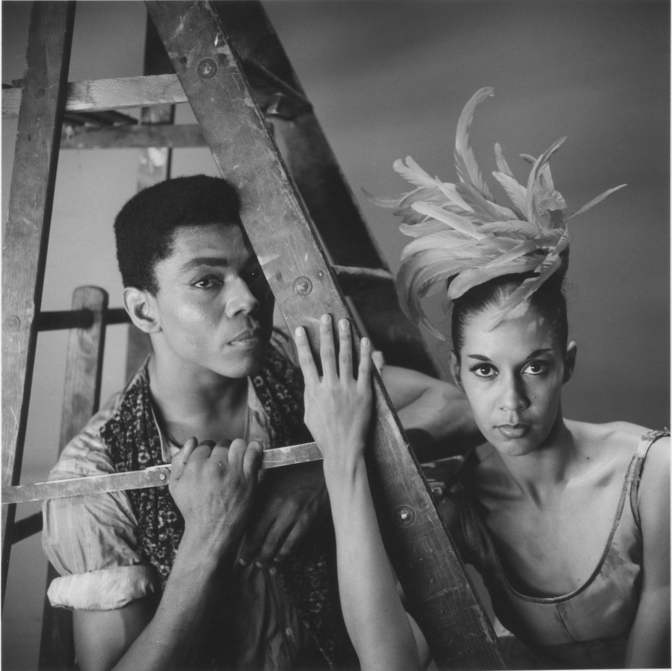 A black-and-white image of Alvin Ailey and Carmen DeLavallade in costume, zoomed in showing their torsos and faces. Ailey poses under a ladder.