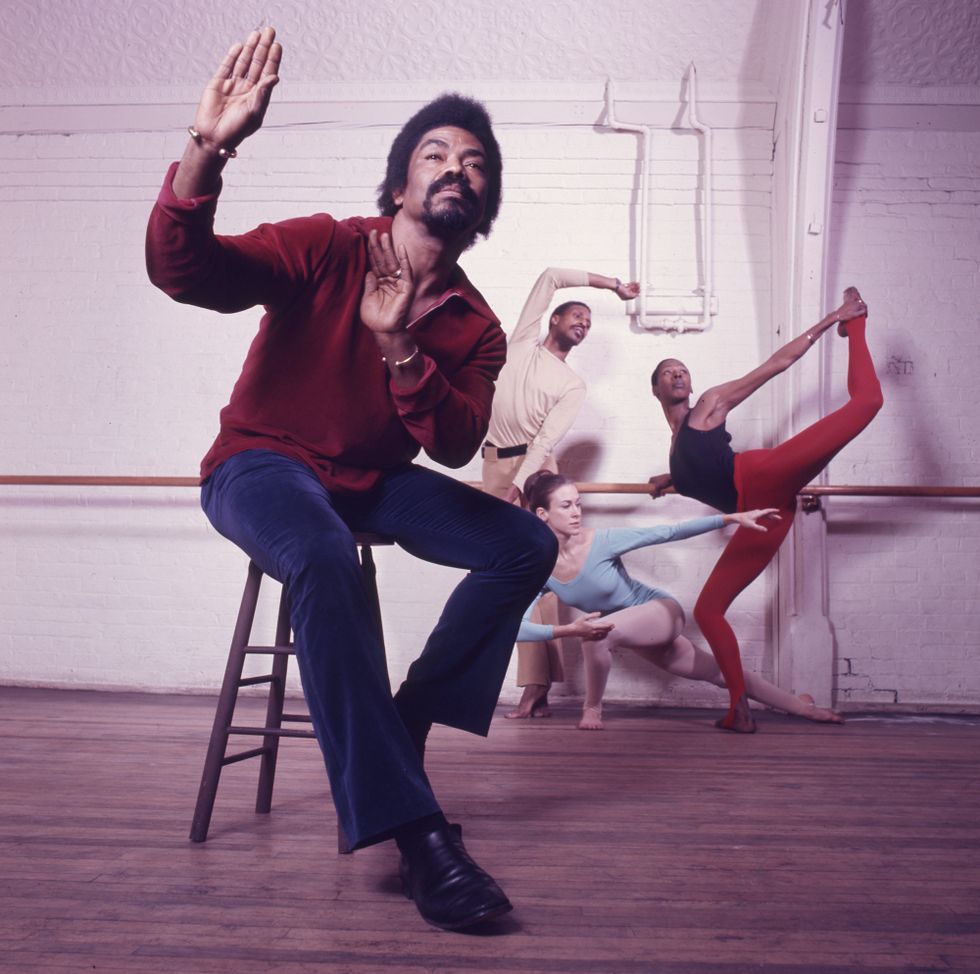 A color photo in a dance studio. Alvin Ailey sits on a stool downstage, while three dancers pose on different levels against the wall and barre in the background.