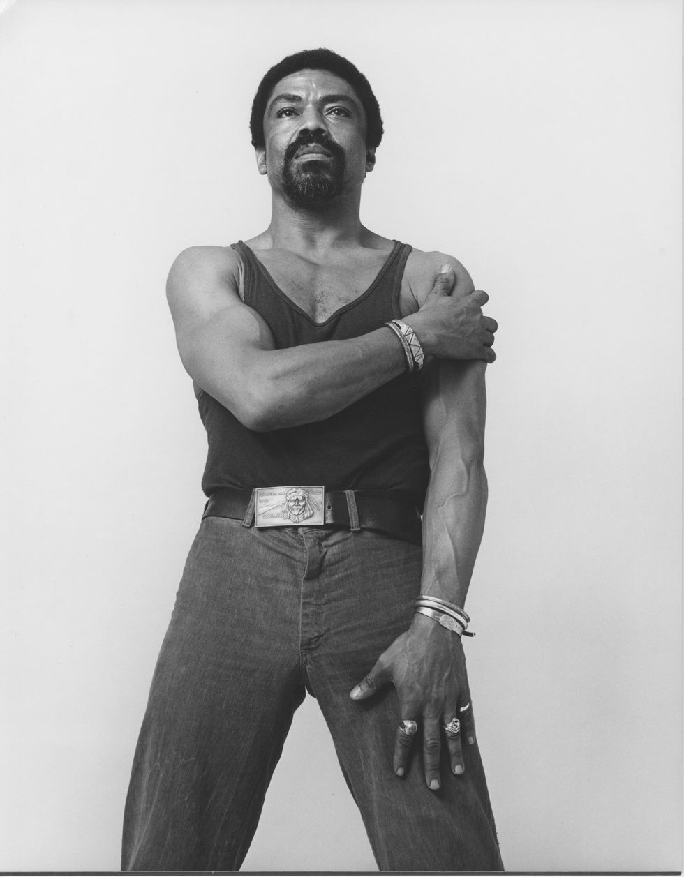 A black-and-white image of Alvin Ailey looking at the camera. he is wearing jeans and a tank top.