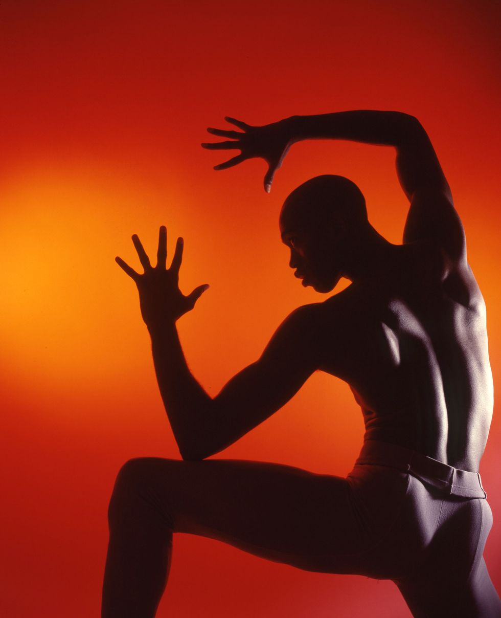 A male dancer faces backwards with knees and arms bent. His silhouette is seen against an orange background.