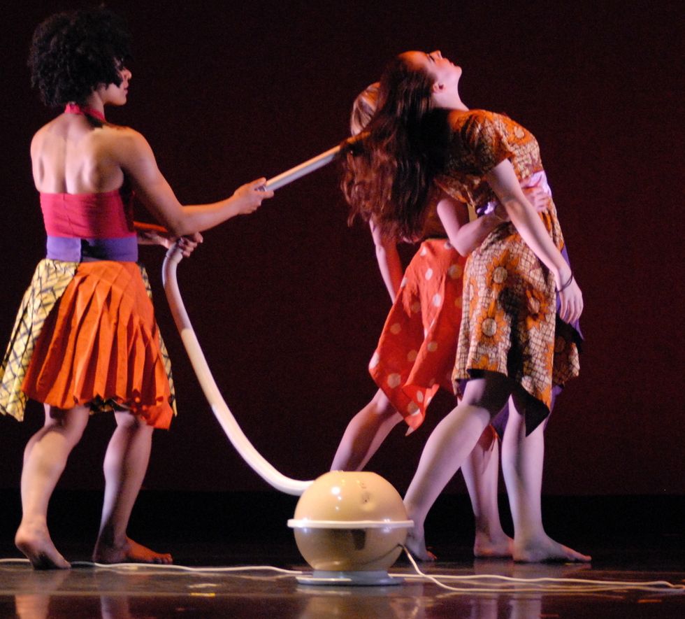 Three women are in brightly colored costumes that look like streetwear. One holds a vaccum with a long extension, and seems to be sucking another dancer's hair with the vaccum. That dancer is walking away from the woman with the vaccum, with the third dancer walking beside and upstage of her