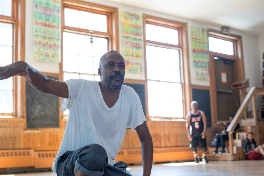 Ozzie Jones sits in a dance studio, his right arm lifted, wearing jeans and a white t-shirt.