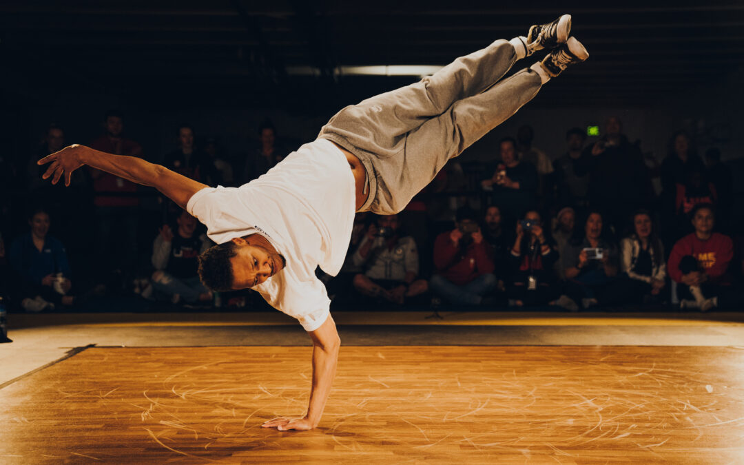 Morris "Bboy Morris" Isby balances on one arm, his legs both extended to 2 o'clock while his head is at 8 o'clock. He wears a white t-shirt, grey sweats, and well worn black Converse. Many members of the crowd watching at the side of a wooden dance floor have expressions of shock and delight.