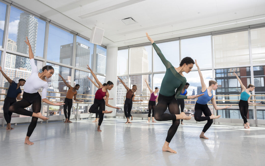 a large group of dancers in a studio with big windows, all posed with leg crossed and lateral bend