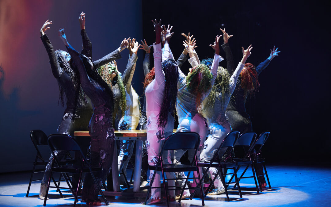 a group of dancers surrounding a table standing out of their chairs with arms lifted overhead