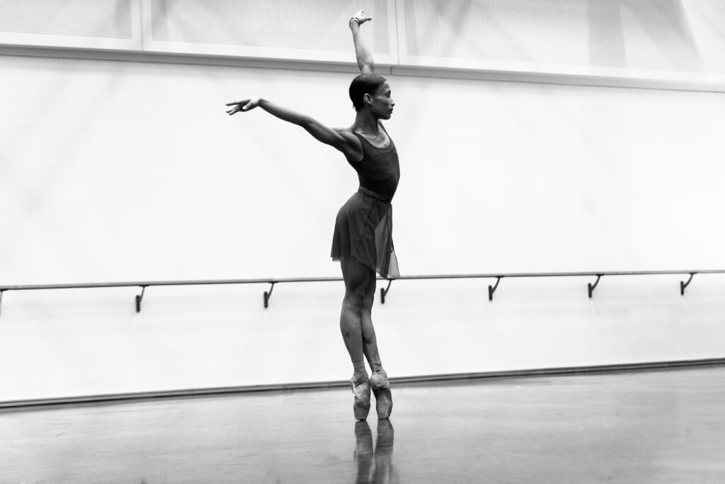 a dancer en pointe performing a tight sous sous with both arms up in a large studio