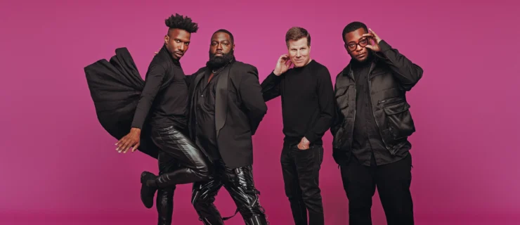 Four men dressed all in black pose against a magenta backdrop. Omari Wiles balances on one leg as his cape flies up behind him, while Arturo Lyons leans into him, one knee bent and the other leg extended long in front. Bill Rauch, the only white man in the group, smiles a little as he brings a hand to the side of his face. To his left, Zhailon Levingston looks seriously at the camera, adjusting his black rimmed glasses with one hand.