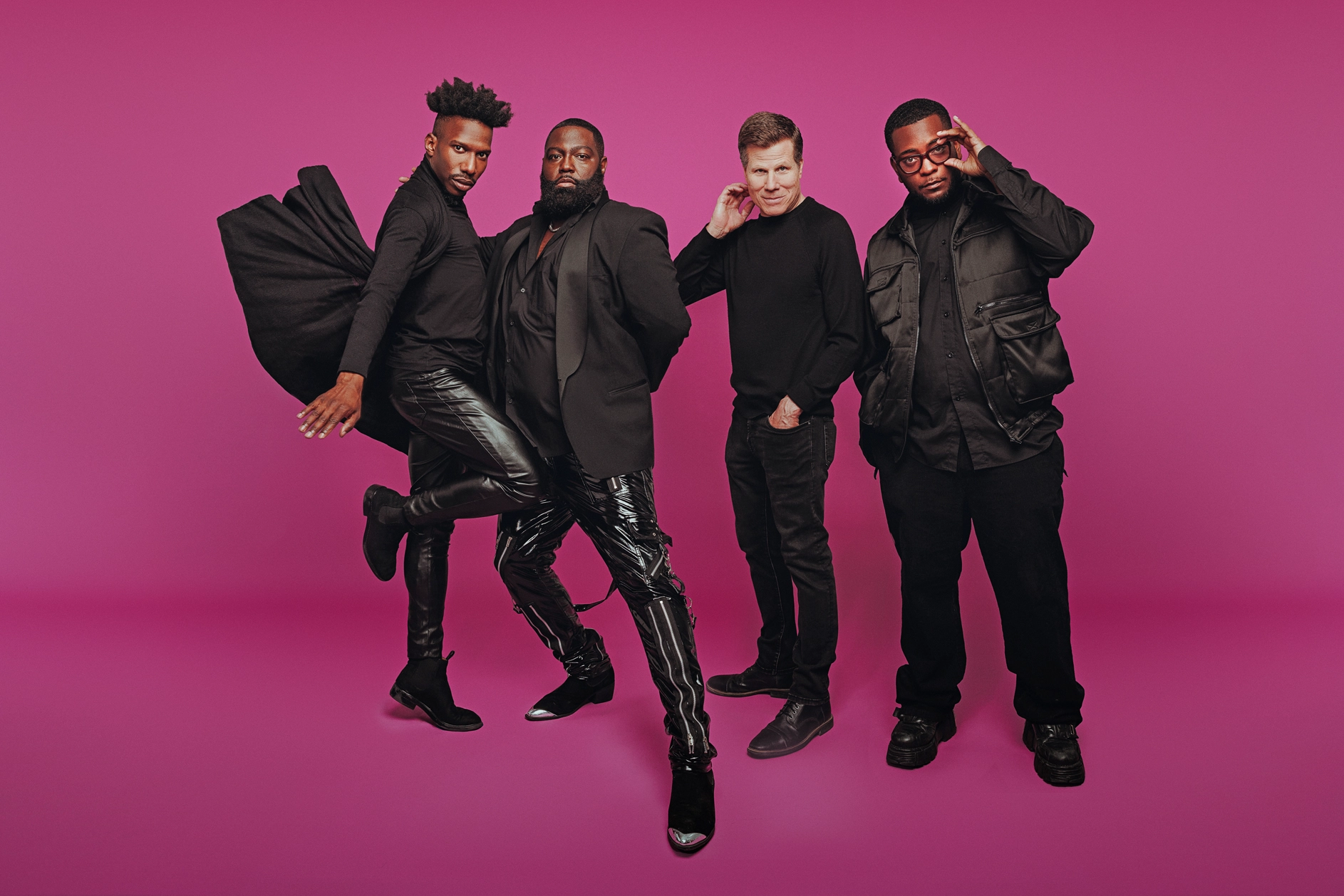 Four men dressed all in black pose against a magenta backdrop. Omari Wiles balances on one leg as his cape flies up behind him, while Arturo Lyons leans into him, one knee bent and the other leg extended long in front. Bill Rauch, the only white man in the group, smiles a little as he brings a hand to the side of his face. To his left, Zhailon Levingston looks seriously at the camera, adjusting his black rimmed glasses with one hand.