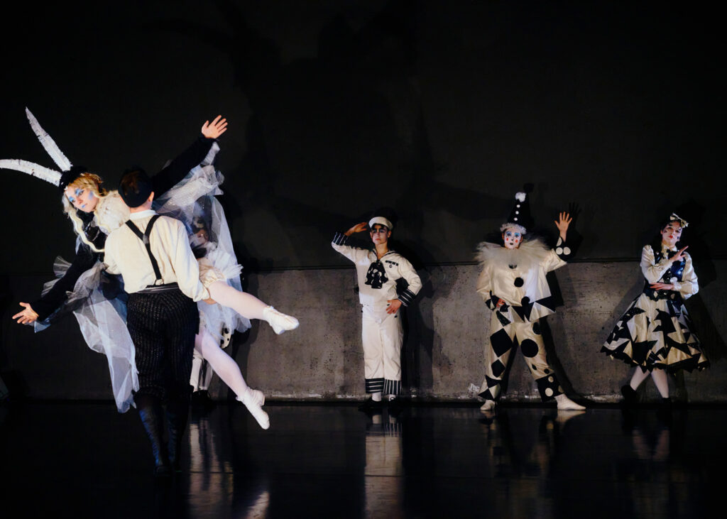 Five dancers in graphic black-and-white costumes and makeup perform in front of a black backdrop.
