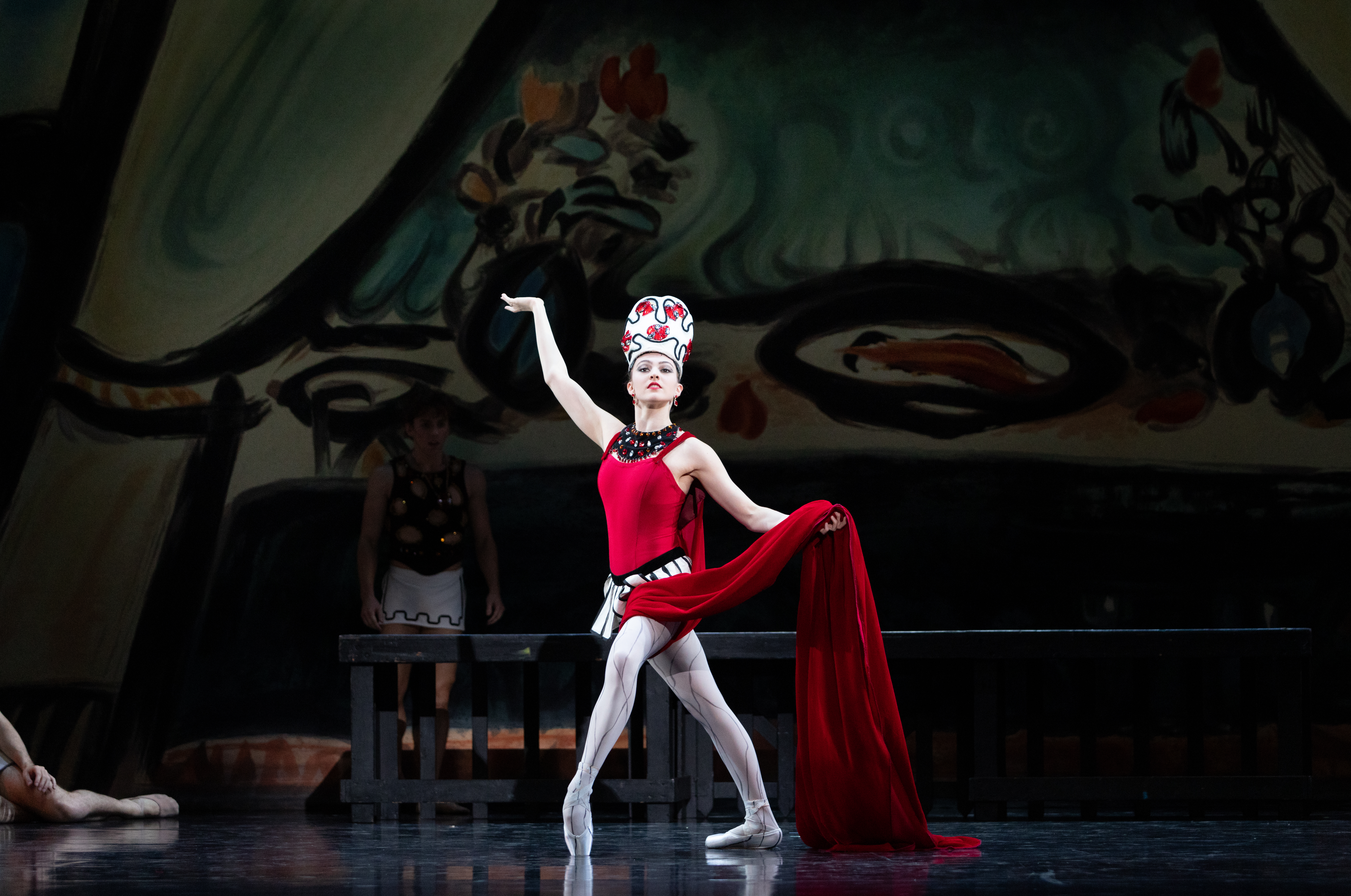 Sydney Dolan onstage as the Siren in Balanchine's Prodigal Son. She poses center stage in a fourth position lunge, front foot in forced arch on pointe. Her left hand holds the long red train of her costume at waist height; her right hand is raised to the side, palm up.
