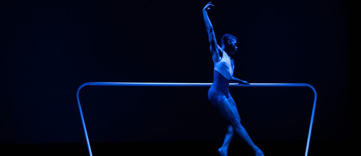 A lone dancer stands at a blue-lit barre. They are in forced arch as they tendu forward, upper arm in an exaggerated high fifth while the other rests lightly on the barre.