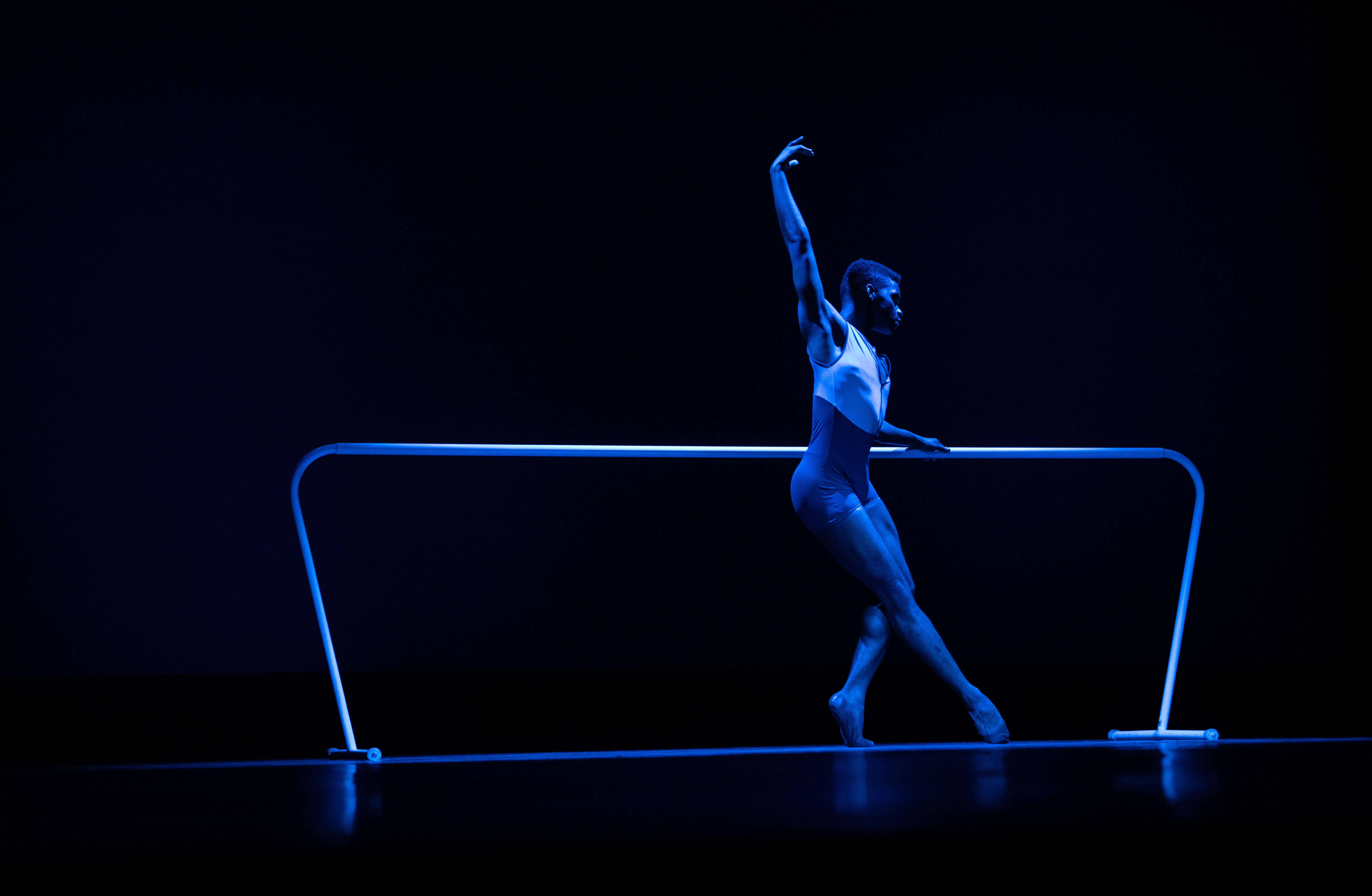 A lone dancer stands at a blue-lit barre. They are in forced arch as they tendu forward, upper arm in an exaggerated high fifth while the other rests lightly on the barre.