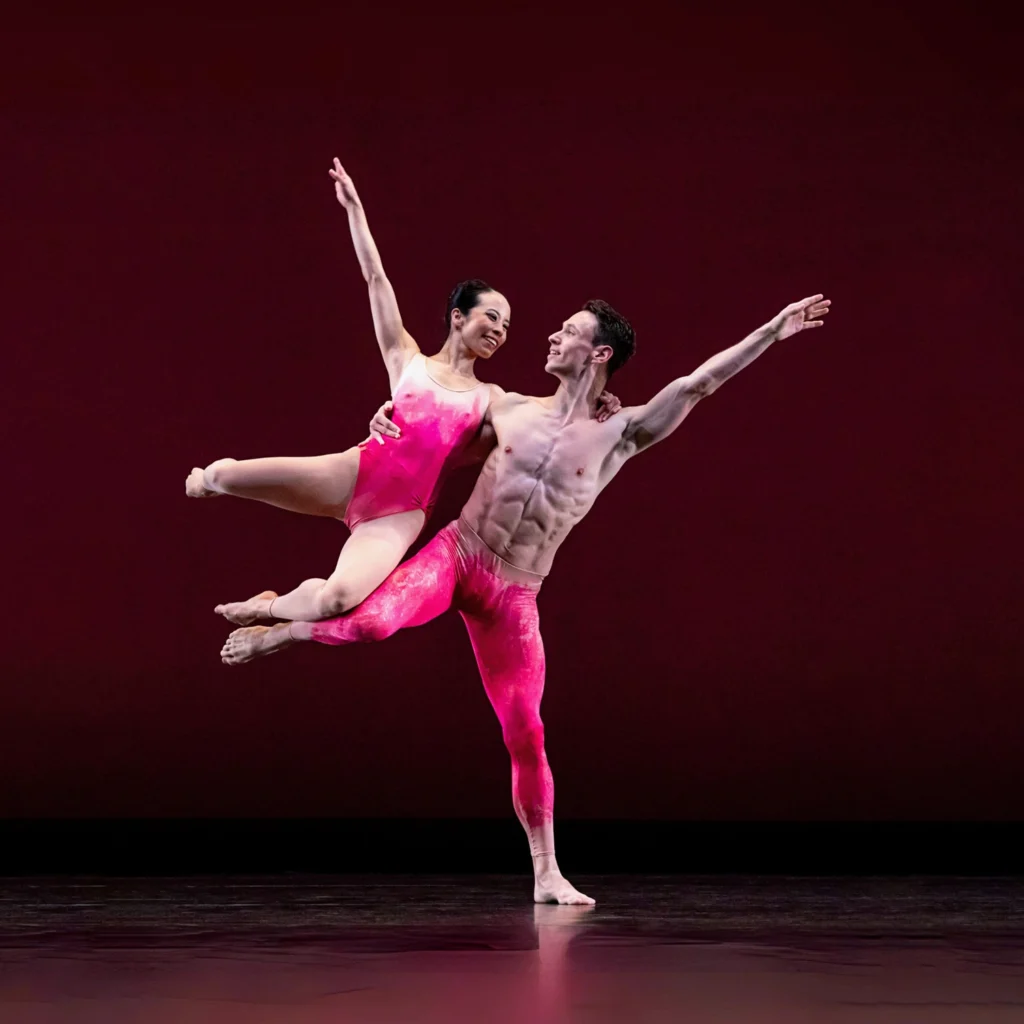 Two dancers smile brightly at each other mid-flight. The shirtless male dancer balances on one leg, his working leg raised in attitude side. A leotard-wearing female dancer flies through the air, both knees bent as she appears to rest on her partner's upraised leg. Their inside arms wrap around each other's shoulders, while their outside arms rise at the same angle to the side.