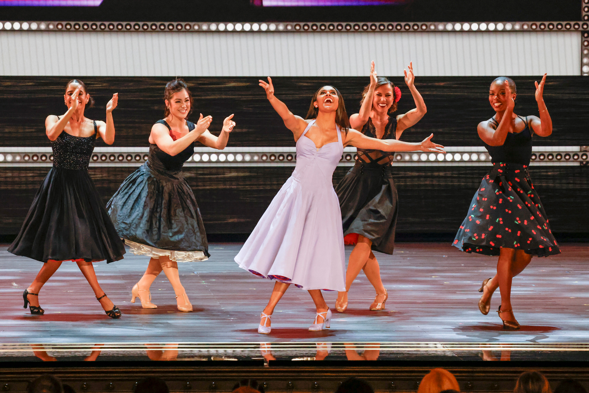DeBose, in a swirly lilac dress, poses onstage in front of four other clapping dancers in similarly full-skirted black dresses.
