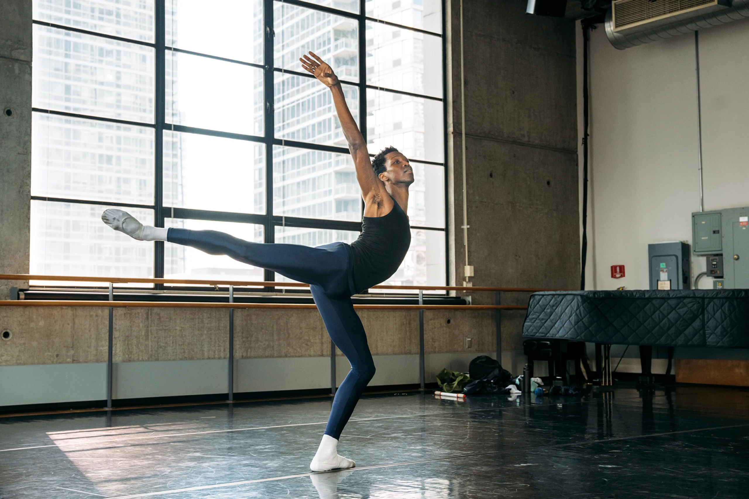 Calvin Royal III poses in arabesque, standing leg in plié and working side arm in a high alongé. He is in a studio with large windows and dark marley; a piano sits in the back corner.