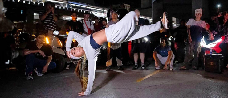 Logan "Logistx" Edra in a cypher. She is captured balancing on one hand, face up, body parallel to the floor. Her other hand touches her head; one leg bends over top the other as it kicks out straight.