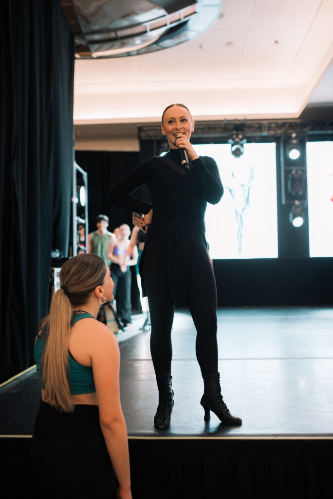 a teacher dressed in all black wearing heels talking into a microphone on stage