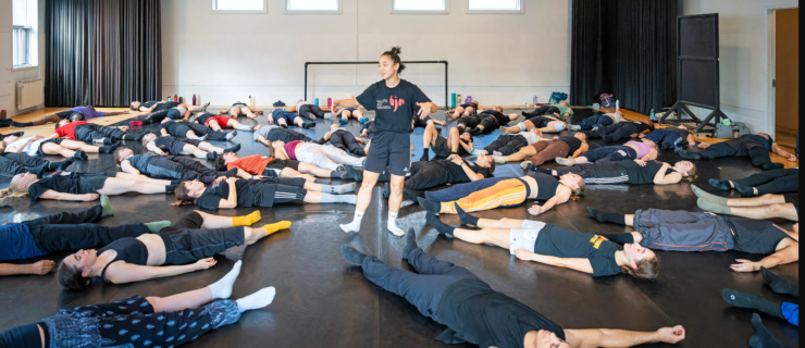 an instructor standing in the middle of a studio with students lying on their backs on the floor