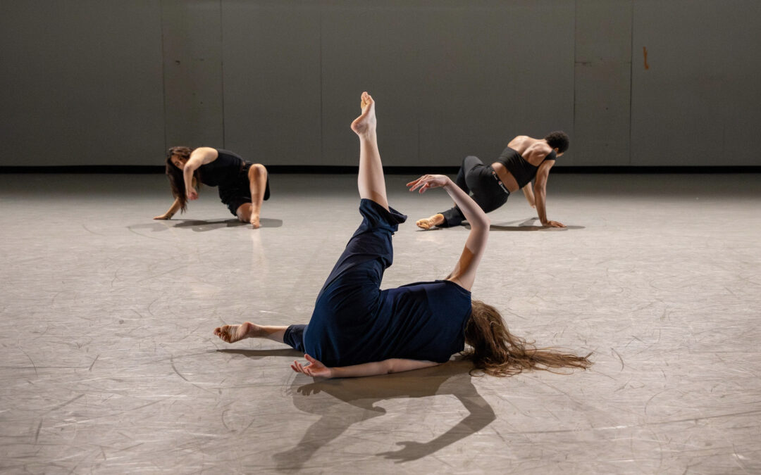 Three dancers in dark-colored practice clothes are caught mid-movement on the gray floor of a dance studio.