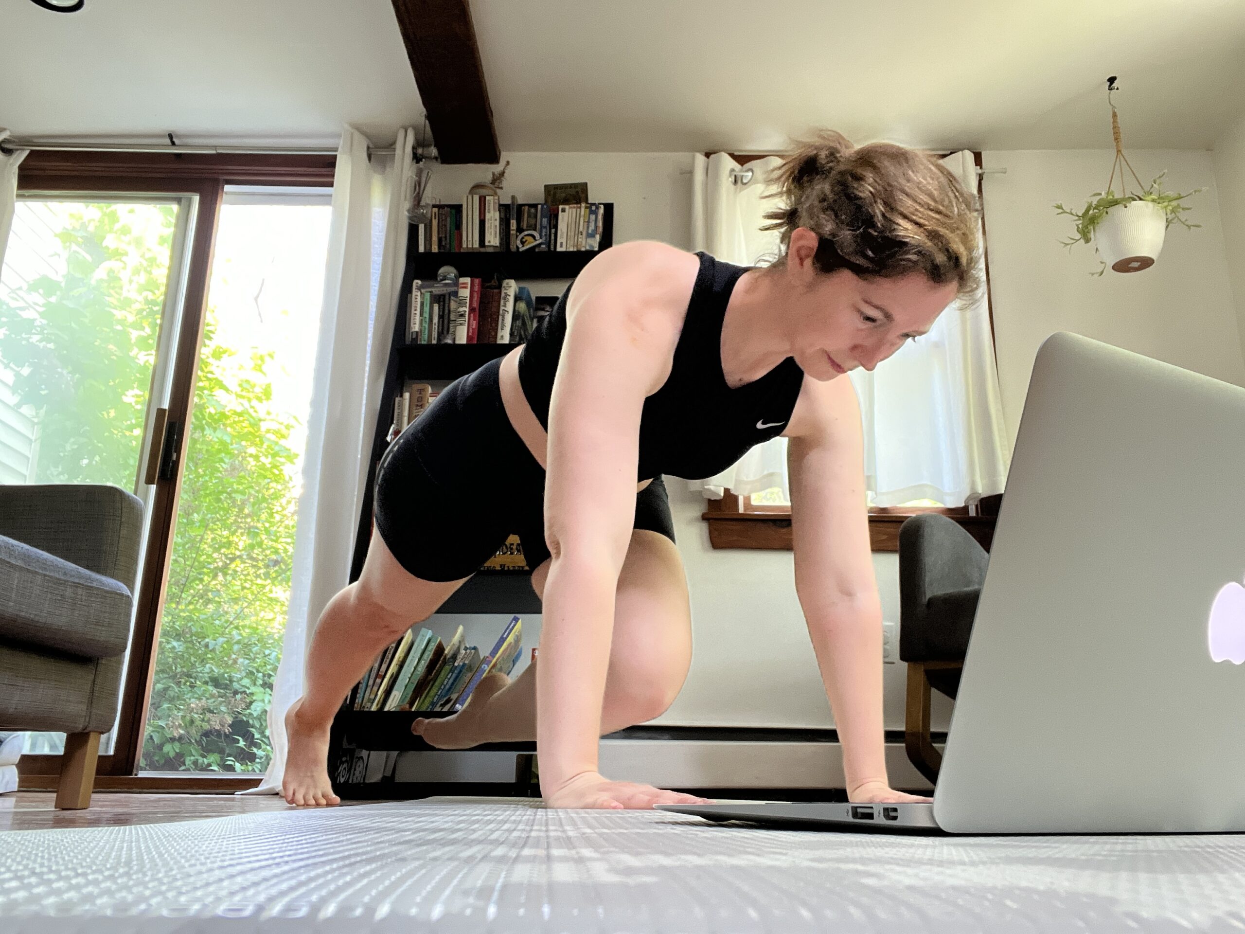 Heimlich, wearing a black sports bra and bike shorts, is seen in her living room with her laptop on the floor in front of her. She looks at the screen as she executes a modified plank position, with her left leg raised in a parallel passé.