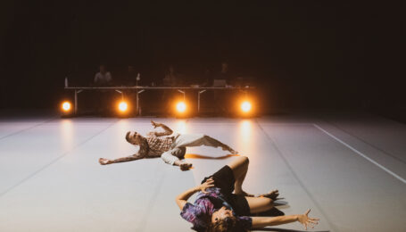 Obergfell and Delorme writhe on a gray dance floor, illuminated by four bright floor lights. In the blurred background, four shadowy figures sit at a table strewn with wires and laptops.