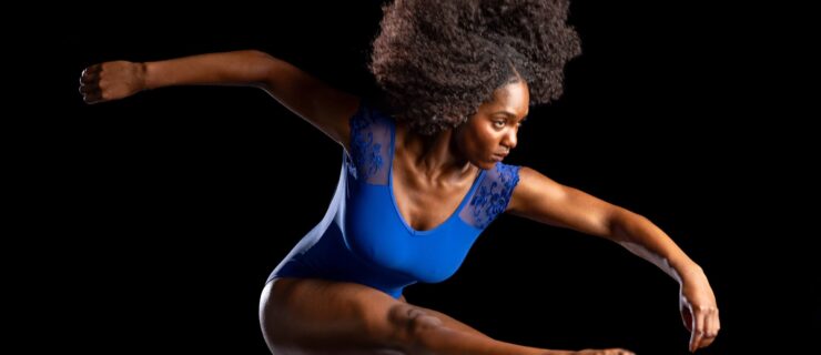 a female dancer wearing a blue leotard jumping in the air against a dark backdrop