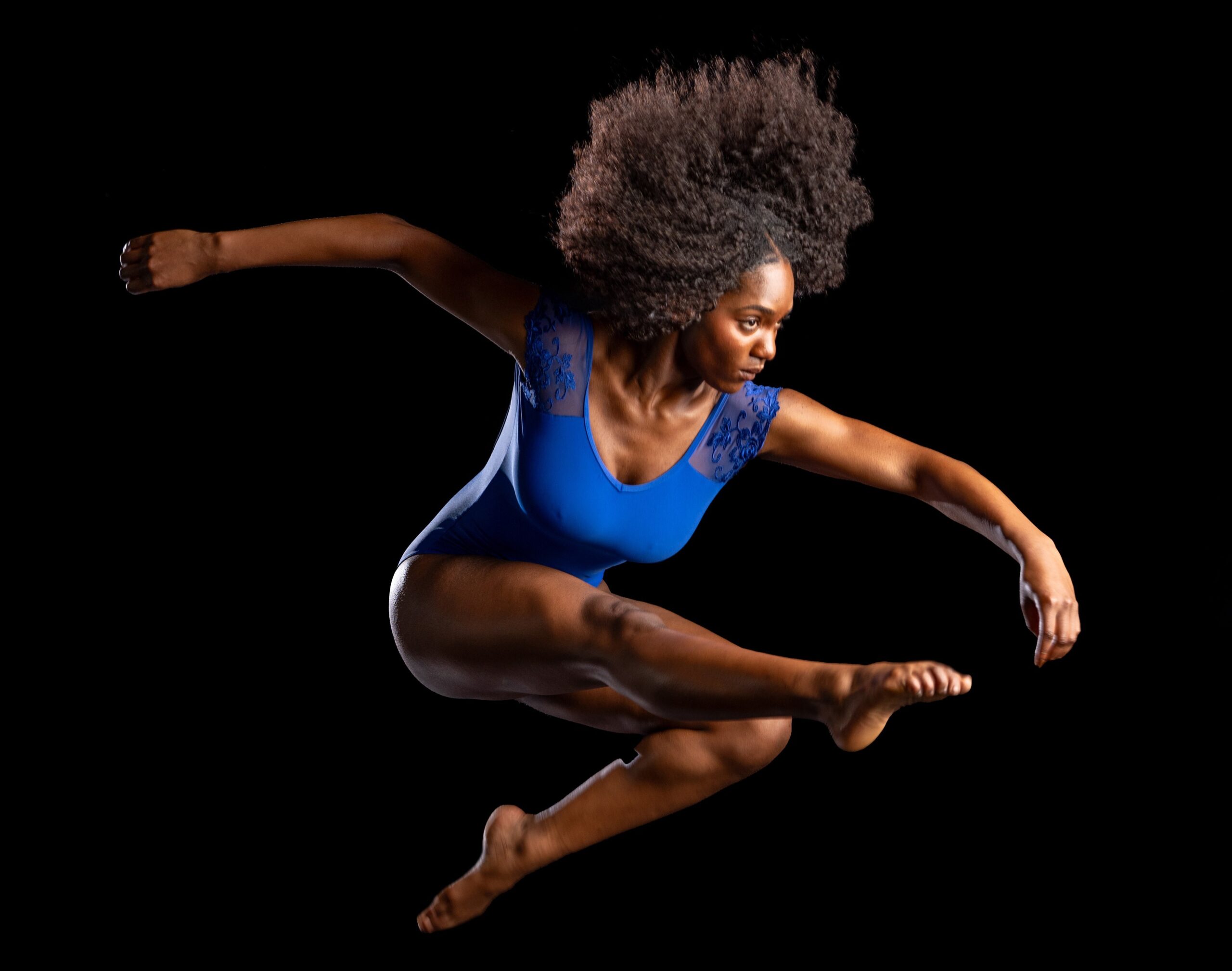 a female dancer wearing a blue leotard jumping in the air against a dark backdrop