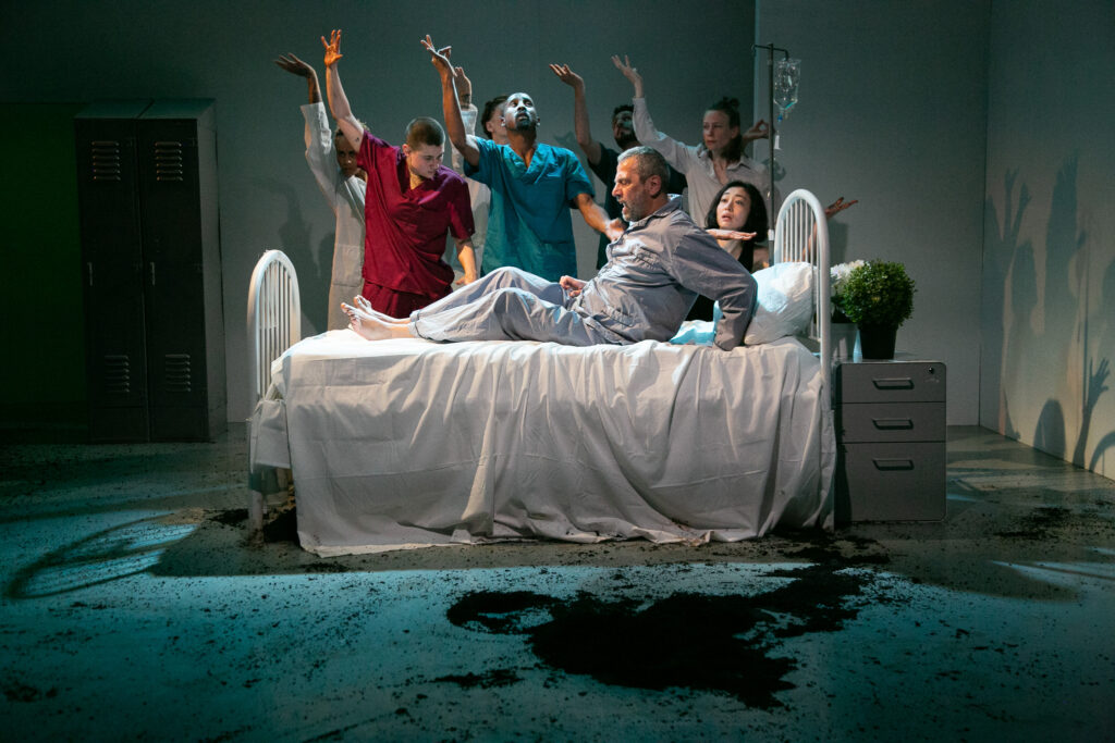 A man in pajamas startles upright from where he is lying on a white bed. A half dozen dancers are upstage of the bed, right arms bent and raised overhead, palms tipping up. Another dancer looks curiously at the first man, hands tucked beneath their chin as they lean on the bed. Downstage, something dark splatters the floor beside the bed.