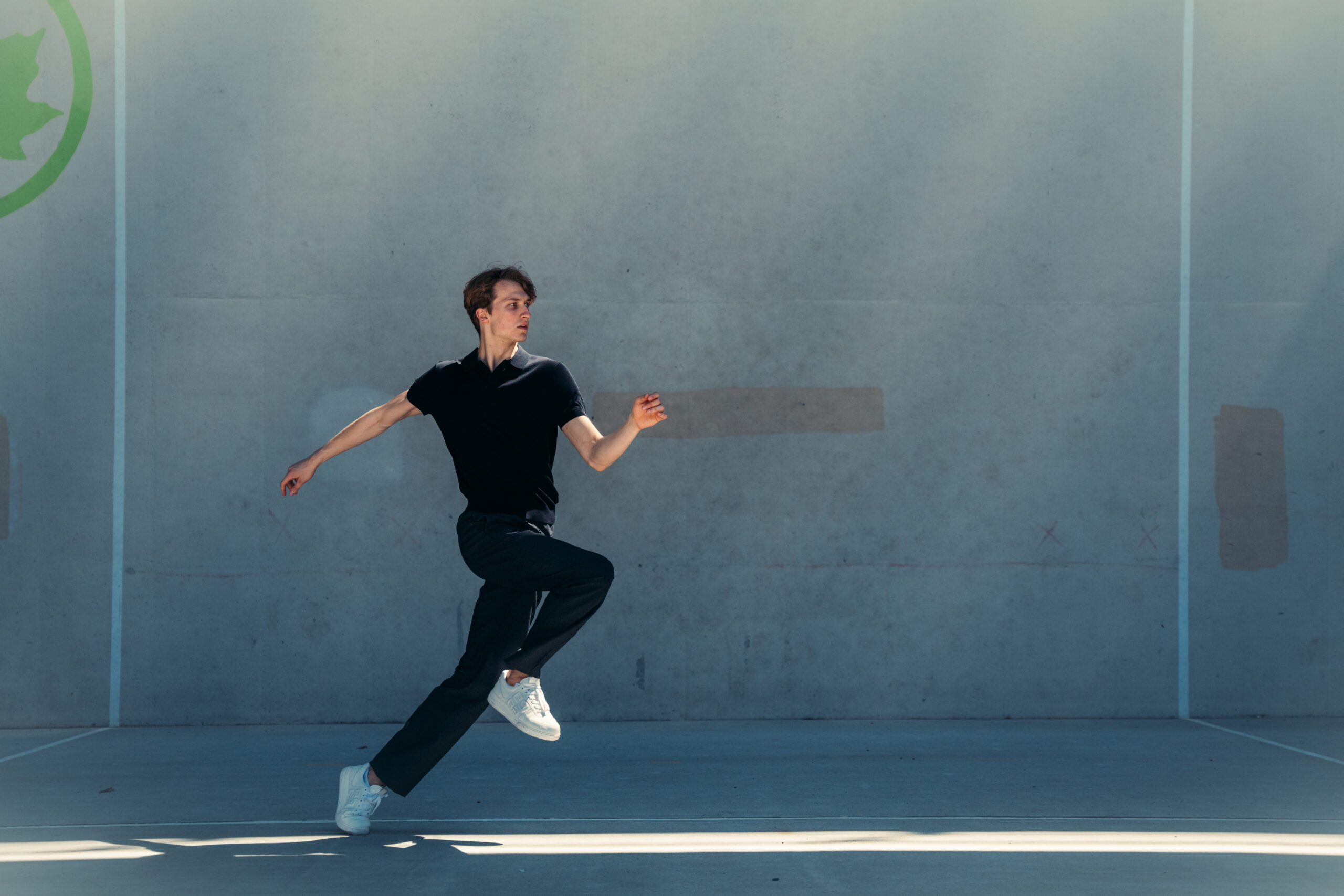 a male dancer pushing off one leg with the other lifted in front while dancing outside by a large wall